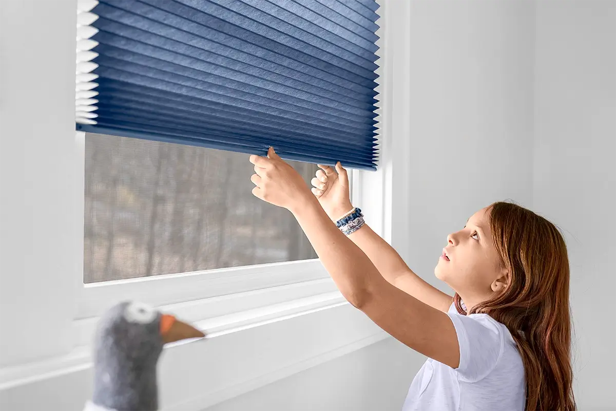 A young girl reaches up to pull down blue cellular shades