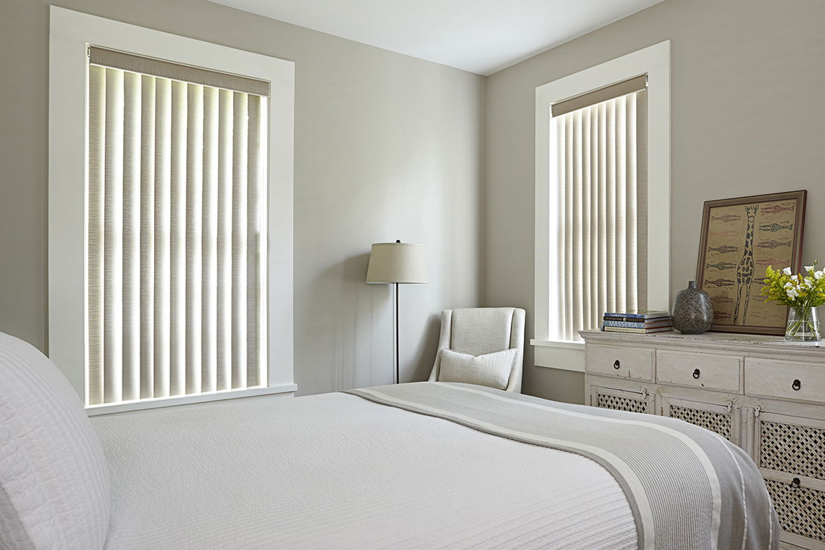 Cream-colored vinyl vertical blinds cover two windows in a modern bedroom.