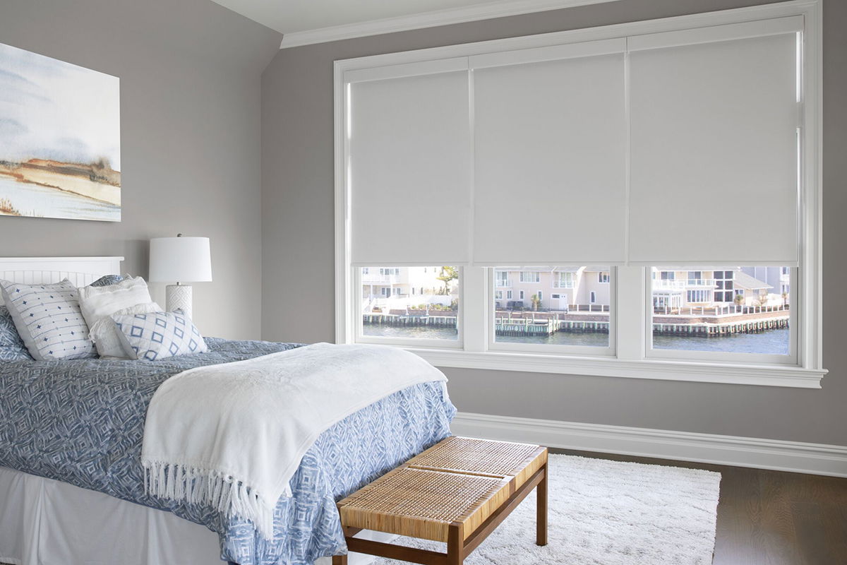 Blackout white roller shades cover three side-by-side windows in a modern waterfront bedroom